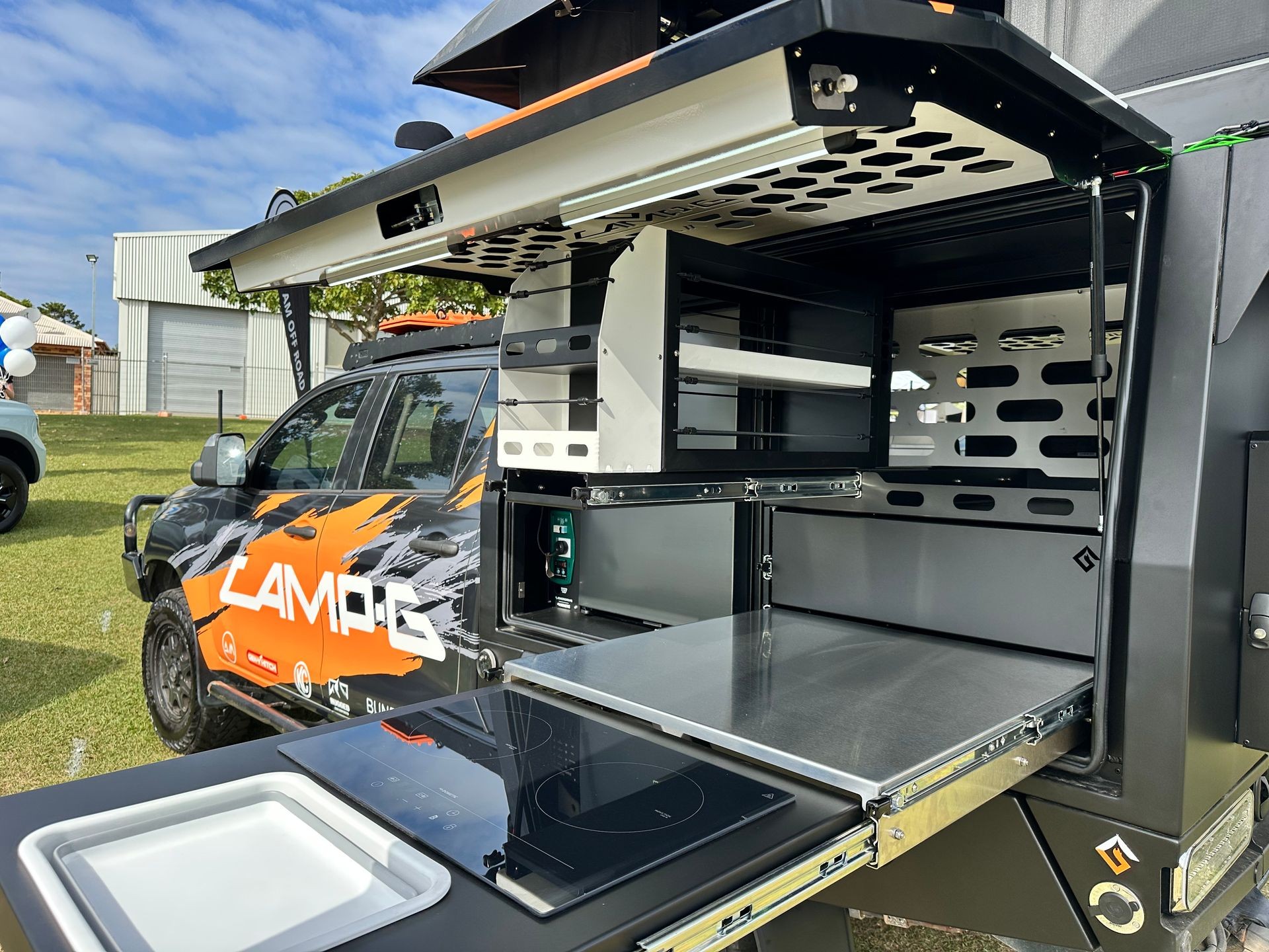 Camper setup with open compartments and cooking area next to a decorated off-road vehicle on grass.